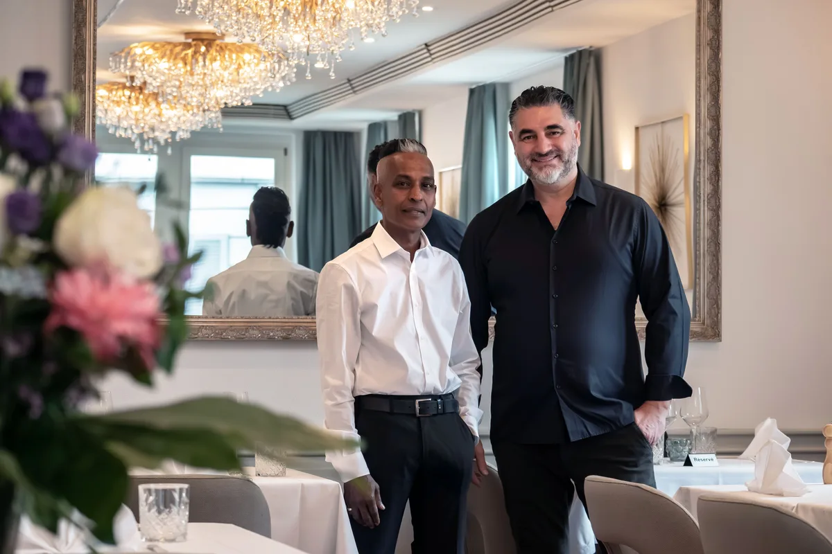 Fabio Botta und sein Kellner Filippo posieren im Restaurant. Fabio trägt ein schwarzes Hemd, der deutlich kleinere, schmächtige Kellner ein weisses Hemd und schwarze Hosen.